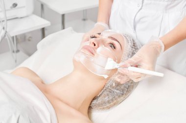 Kozmetik uzmanı kadın yüzüne cilt tazeleme için krem maskesi, güzellik salonundaki yaşlanma karşıtı kozmetik prosedürleri uyguluyor. Eldivenli güzellik uzmanı elleri kozmetik fırça tutuyor ve kozmetik maske takıyor