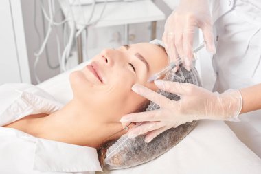 Kozmetik uzmanı kadın tenine gençleştirme enjeksiyonu yapıyor güzellik salonundaki yaşlanma karşıtı kozmetik prosedürü. Eldivenli güzellik uzmanı yüz asidi enjeksiyonu tedavisi yapıyor.