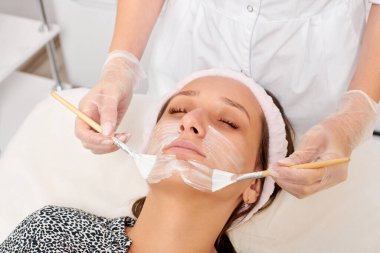 Kozmetik uzmanı, güzellik salonunda cildini yenilemek için kadın yüzüne kozmetik krem maskesi uyguluyor. Eldivenli güzellik uzmanı iki fırçalı kozmetik krem maskesi takıyor.