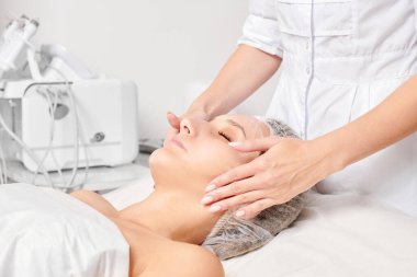 Güzellik salonunda kozmetik uzmanı cildine nemlendirici krem maskeyi sürüp gençleştirme, yaşlanmayı önleyici kozmetik prosedür uyguluyor. Güzellik uzmanı elleri kadın derisi için yüz kremi sürüyor.