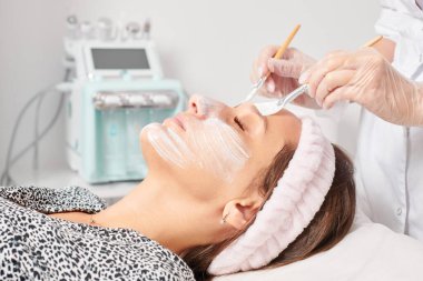 Güzellik uzmanı, güzellik salonunda estetik krem maskesini cilt tazeleme, nemlendirici kozmetik işlemleri için kadın yüzüne uyguluyor. Eldivenli kozmetik uzmanı iki fırçayla kozmetik krem maskesi takıyor.