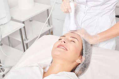 Kozmetik uzmanı, güzellik salonundaki gençleşen kadın yüzü için RF kaldırma, yaşlanma karşıtı kozmetik prosedürü yapıyor. Güzellik uzmanı kadın yüz derisinde artan kolajen için radyo frekansı tedavisi uyguluyor.