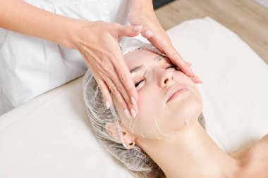 Güzellik uzmanı, güzellik salonundaki cilt bakımı için kadın yüzüne çarşaf maskesi, kırışıklık önleyici kozmetik prosedürler takıyor. Kozmetik uzmanı kadın yüzüne nemlendirici çarşaf maskesi uyguluyor.