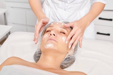 Güzellik salonunda kozmetik uzmanı cildine nemlendirici krem maskeyi sürüp gençleştirme, yaşlanmayı önleyici kozmetik prosedür uyguluyor. Güzellik uzmanı elleri kadın derisi için yüz kremi sürüyor.