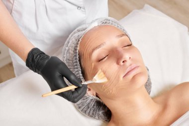 Kozmetik uzmanı kadın yüzüne nemlendirici yüz derisi ve güzellik salonundaki yaşlanma karşıtı kozmetik prosedürü için bal maskesi takıyor. Siyah eldivenli güzellik uzmanı, bal maskesi takmak için kozmetik fırça tutuyor.