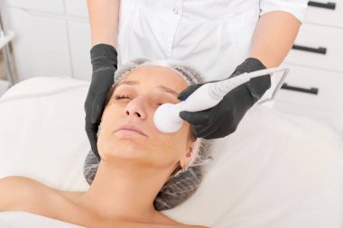 Kozmetik uzmanı, güzellik salonundaki gençleşen kadın yüzü için RF kaldırma, yaşlanma karşıtı kozmetik prosedürü yapıyor. Güzellik uzmanı kadın yüz derisinde artan kolajen için radyo frekansı tedavisi uyguluyor.