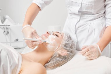 Kozmetik uzmanı kadın yüzüne cilt tazeleme için krem maskesi, güzellik salonundaki yaşlanma karşıtı kozmetik prosedürleri uyguluyor. Eldivenli güzellik uzmanı elleri kozmetik fırça tutuyor ve kozmetik maske takıyor