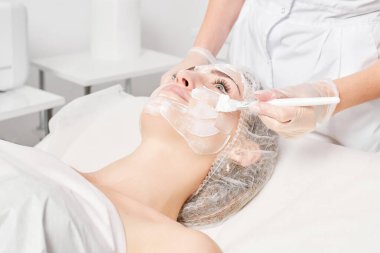 Güzellik uzmanı, güzellik salonlarında cilt bakımı için kadın yüzüne vitamin kremi maskesi, yaşlanma karşıtı kozmetik işlemler uyguluyor. Eldivenli kozmetik uzmanı maske takmak için kozmetik fırça tutuyor