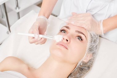 Güzellik uzmanı, güzellik salonlarında cilt bakımı için kadın yüzüne vitamin kremi maskesi, yaşlanma karşıtı kozmetik işlemler uyguluyor. Eldivenli kozmetik uzmanı maske takmak için kozmetik fırça tutuyor