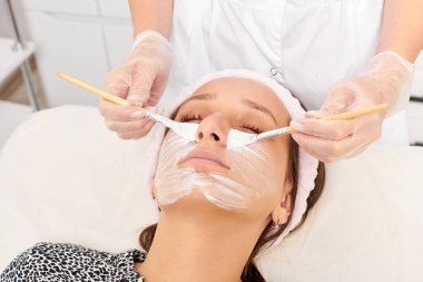 Kozmetik uzmanı, güzellik salonunda cildini yenilemek için kadın yüzüne kozmetik krem maskesi uyguluyor. Eldivenli güzellik uzmanı iki fırçalı kozmetik krem maskesi takıyor.