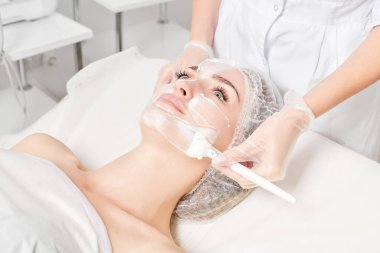 Güzellik uzmanı, güzellik salonlarında cilt bakımı için kadın yüzüne vitamin kremi maskesi, yaşlanma karşıtı kozmetik işlemler uyguluyor. Eldivenli kozmetik uzmanı maske takmak için kozmetik fırça tutuyor