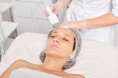 Kozmetik uzmanı, güzellik salonundaki yaşlanma karşıtı kozmetik prosedürleri, cilt bakımını yenilemek için yüz ultrasoniği yapıyor. Güzellik uzmanı yüz bakımını ultrason fırçasıyla yapıyor.