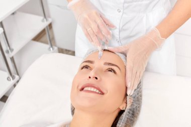 Kozmetik uzmanı kadın tenine gençleştirme enjeksiyonu yapıyor güzellik salonundaki yaşlanma karşıtı kozmetik prosedürü. Eldivenli güzellik uzmanı yüz asidi enjeksiyonu tedavisi yapıyor.