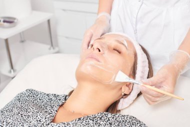 Kozmetik uzmanı, güzellik salonundaki kozmetik bakım ürünlerini nemlendirmek için kadın yüzüne estetik krem takıyor. Eldivenli güzellik uzmanı fırçayla kozmetik krem maskesi takıyor.