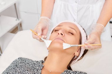 Kozmetik uzmanı, güzellik salonunda cildini yenilemek için kadın yüzüne kozmetik krem maskesi uyguluyor. Eldivenli güzellik uzmanı iki fırçalı kozmetik krem maskesi takıyor.