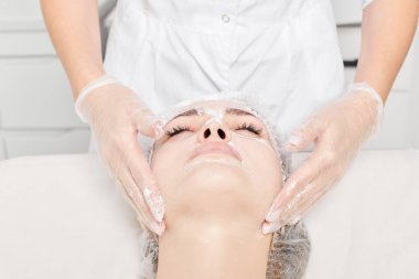 Kozmetik uzmanı, güzellik salonundaki yaşlanma karşıtı kozmetik prosedürleri ve cilt bakımı için kadın yüzüne krem masajı yapıyor. Eldivenli güzellik uzmanı elleri kadın yüzüne krem sürüyor.