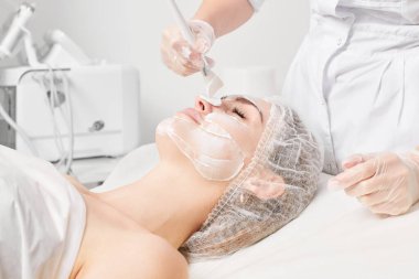Güzellik uzmanı, güzellik salonlarında cilt bakımı için kadın yüzüne vitamin kremi maskesi, yaşlanma karşıtı kozmetik işlemler uyguluyor. Eldivenli kozmetik uzmanı maske takmak için kozmetik fırça tutuyor