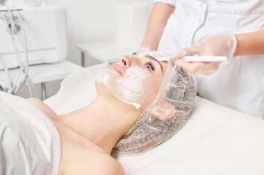 Güzellik uzmanı, güzellik salonlarında cilt bakımı için kadın yüzüne vitamin kremi maskesi, yaşlanma karşıtı kozmetik işlemler uyguluyor. Eldivenli kozmetik uzmanı maske takmak için kozmetik fırça tutuyor