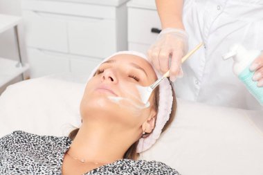 Kozmetik uzmanı, güzellik salonundaki kozmetik bakım ürünlerini nemlendirmek için kadın yüzüne estetik krem takıyor. Eldivenli güzellik uzmanı fırçayla kozmetik krem maskesi takıyor.