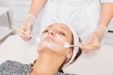 Kozmetik uzmanı, güzellik salonunda cildini yenilemek için kadın yüzüne kozmetik krem maskesi uyguluyor. Eldivenli güzellik uzmanı iki fırçalı kozmetik krem maskesi takıyor.