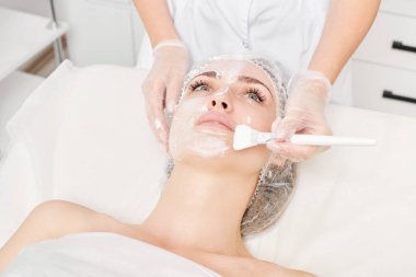 Güzellik uzmanı, güzellik salonlarında cilt bakımı için kadın yüzüne vitamin kremi maskesi, yaşlanma karşıtı kozmetik işlemler uyguluyor. Eldivenli kozmetik uzmanı maske takmak için kozmetik fırça tutuyor
