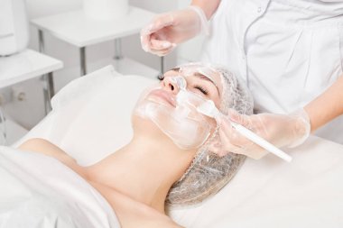 Kozmetik uzmanı kadın yüzüne cilt tazeleme için krem maskesi, güzellik salonundaki yaşlanma karşıtı kozmetik prosedürleri uyguluyor. Eldivenli güzellik uzmanı elleri kozmetik fırça tutuyor ve kozmetik maske takıyor