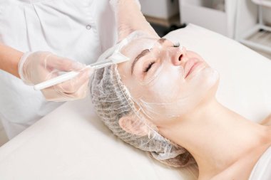 Güzellik uzmanı, güzellik salonundaki yaşlanma karşıtı kozmetik işlemler için cildini yenilemek için kadın yüzüne kozmetik krem maskesi sürüyor. Eldivenleri teslim eden kozmetik uzmanı maske takmak için kozmetik fırça tutuyor.