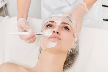 Kozmetik uzmanı kadın yüzüne cilt tazeleme için krem maskesi, güzellik salonundaki yaşlanma karşıtı kozmetik prosedürleri uyguluyor. Eldivenli güzellik uzmanı elleri kozmetik fırça tutuyor ve kozmetik maske takıyor