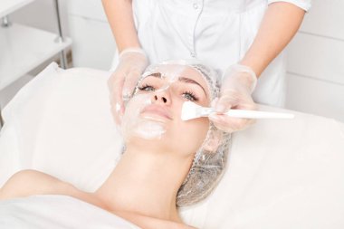 Güzellik uzmanı, güzellik salonundaki yaşlanma karşıtı kozmetik işlemler için cildini yenilemek için kadın yüzüne kozmetik krem maskesi sürüyor. Eldivenleri teslim eden kozmetik uzmanı maske takmak için kozmetik fırça tutuyor.