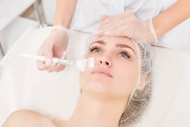 Kozmetik uzmanı kadın yüzüne cilt tazeleme için krem maskesi, güzellik salonundaki yaşlanma karşıtı kozmetik prosedürleri uyguluyor. Eldivenli güzellik uzmanı elleri kozmetik fırça tutuyor ve kozmetik maske takıyor