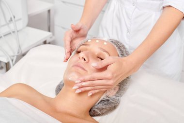 Güzellik salonunda kozmetik uzmanı cildine nemlendirici krem maskeyi sürüp gençleştirme, yaşlanmayı önleyici kozmetik prosedür uyguluyor. Güzellik uzmanı elleri kadın derisi için yüz kremi sürüyor.