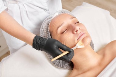 Güzellik uzmanı kadın yüzüne nemlendirici yüz derisi ve güzellik salonundaki yaşlanma karşıtı kozmetik prosedürleri için bal maskesi takıyor. Siyah eldivenli kozmetik uzmanı bal maskesi takmak için kozmetik fırça tutuyor.