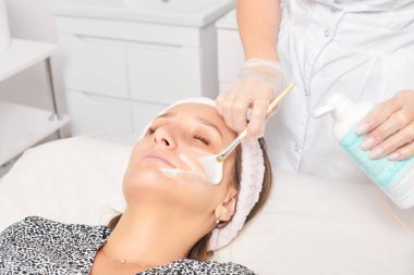 Güzellik uzmanı, güzellik salonundaki kozmetik krem maskesini cilt tazeleme, nemlendirici kozmetik işlemler için kadın yüzüne uyguluyor. Eldivenli kozmetik uzmanı fırçayla kozmetik krem maskesi takıyor.