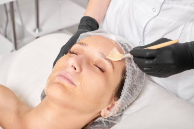 Güzellik uzmanı kadın yüzüne nemlendirici yüz derisi ve güzellik salonundaki yaşlanma karşıtı kozmetik prosedürleri için bal maskesi takıyor. Siyah eldivenli kozmetik uzmanı bal maskesi takmak için kozmetik fırça tutuyor.