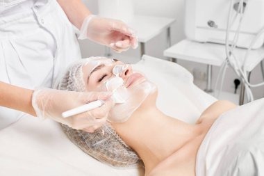 Kozmetik uzmanı kadın yüzüne cilt tazeleme için krem maskesi, güzellik salonundaki yaşlanma karşıtı kozmetik prosedürleri uyguluyor. Eldivenli güzellik uzmanı elleri kozmetik fırça tutuyor ve kozmetik maske takıyor