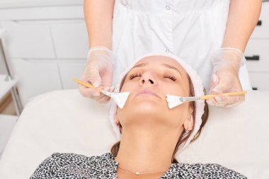 Kozmetik uzmanı, güzellik salonunda cildini yenilemek için kadın yüzüne kozmetik krem maskesi uyguluyor. Eldivenli güzellik uzmanı iki fırçalı kozmetik krem maskesi takıyor.