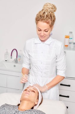 Güzellik uzmanları, güzellik salonundaki estetik ameliyatları nemlendiren cilt yenileme için kadın yüzüne kozmetik krem maskesi uyguluyor. Eldivenli kozmetik uzmanı fırçayla kozmetik krem maskesi takıyor.