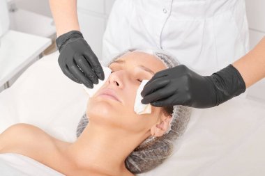 Güzellik uzmanı, güzellik salonundaki yüz kremi maskesini kadın yüzünden kozmetik mendillerle alıyor, cilt yenileme estetik ameliyatı yapıyor. Kozmetik uzmanı yüz maskesini çıkarmak için peçete tutuyor.