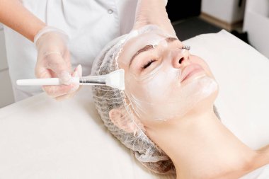 Kozmetik uzmanı kadın yüzüne cilt tazeleme için krem maskesi, güzellik salonundaki yaşlanma karşıtı kozmetik prosedürleri uyguluyor. Eldivenli güzellik uzmanı elleri kozmetik fırça tutuyor ve kozmetik maske takıyor