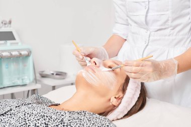 Kozmetik uzmanı, güzellik salonunda cildini yenilemek için kadın yüzüne kozmetik krem maskesi uyguluyor. Eldivenli güzellik uzmanı iki fırçalı kozmetik krem maskesi takıyor.