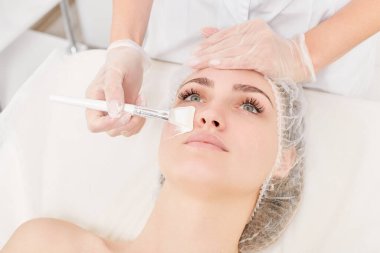 Güzellik uzmanı, güzellik salonundaki yaşlanma karşıtı kozmetik işlemler için cildini yenilemek için kadın yüzüne kozmetik krem maskesi sürüyor. Eldivenleri teslim eden kozmetik uzmanı maske takmak için kozmetik fırça tutuyor.