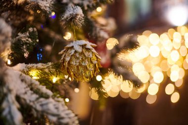 Sarı çelenkli Noel ağacı soğanları, karla kaplı açık hava ksenleri, dekoratif koni lambalı yeşil ağaç, açık hava kış tatili atmosferi. Süslemeleri ve çelenkleri olan Noel ağacı