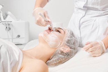 Güzellik uzmanı, güzellik salonlarında cilt bakımı için kadın yüzüne vitamin kremi maskesi, yaşlanma karşıtı kozmetik işlemler uyguluyor. Eldivenli kozmetik uzmanı maske takmak için kozmetik fırça tutuyor