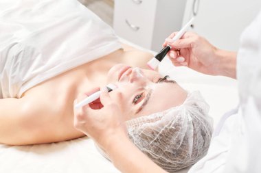 Güzellik salonundaki kozmetik uzmanı cildini tazelemek için kadın yüzüne fırçayla krem sürüyor nemlendirici cilt estetiği uyguluyor. Güzellik uzmanının ellerinde krem sürmek için iki fırça var.