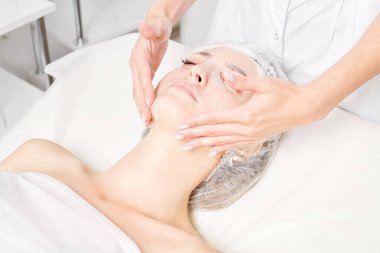 Güzellik uzmanı, güzellik salonundaki cilt bakımı için kadın yüzüne çarşaf maskesi, kırışıklık önleyici kozmetik prosedürler takıyor. Kozmetik uzmanı kadın yüzüne nemlendirici çarşaf maskesi uyguluyor.