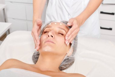 Güzellik uzmanları, güzellik salonlarında gençleştirme için nemlendirici krem maskeyi kadın yüzüne sürerler, yaşlanma karşıtı kozmetik işlemler yaparlar. Kozmetik uzmanı kadın derisi için yüz kremi uyguluyor.