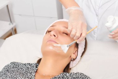 Güzellik uzmanı, güzellik salonundaki kozmetik krem maskesini cilt tazeleme, nemlendirici kozmetik işlemler için kadın yüzüne uyguluyor. Eldivenli kozmetik uzmanı fırçayla kozmetik krem maskesi takıyor.