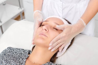 Eldivenli kozmetik uzmanı yenilenme, lenfatik drenaj, güzellik salonundaki krem cilt estetiği için kadın yüz masajı yapıyor. Güzellik uzmanı elleriyle cildini düzleştiriyor.