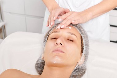 Güzellik salonunda kozmetik uzmanı cildine nemlendirici krem maskeyi sürüp gençleştirme, yaşlanmayı önleyici kozmetik prosedür uyguluyor. Güzellik uzmanı elleri kadın derisi için yüz kremi sürüyor.