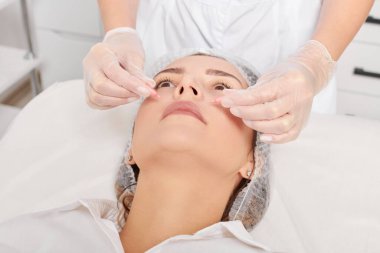 Güzellik uzmanı, güzellik salonundaki yaşlanma karşıtı kozmetik prosedürleri gençleştirme için kadın yüzüne yüz yağı kapsülleri uyguluyor. Eldivenleri teslim eden kozmetolojist beslenme için yağ kapsüllerini tutuyor.